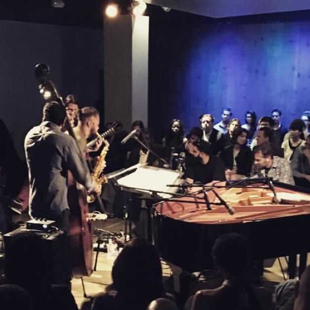 A photo of an intimate jazz club club with dark blue walls. It shows musicians on stage facing an audience.